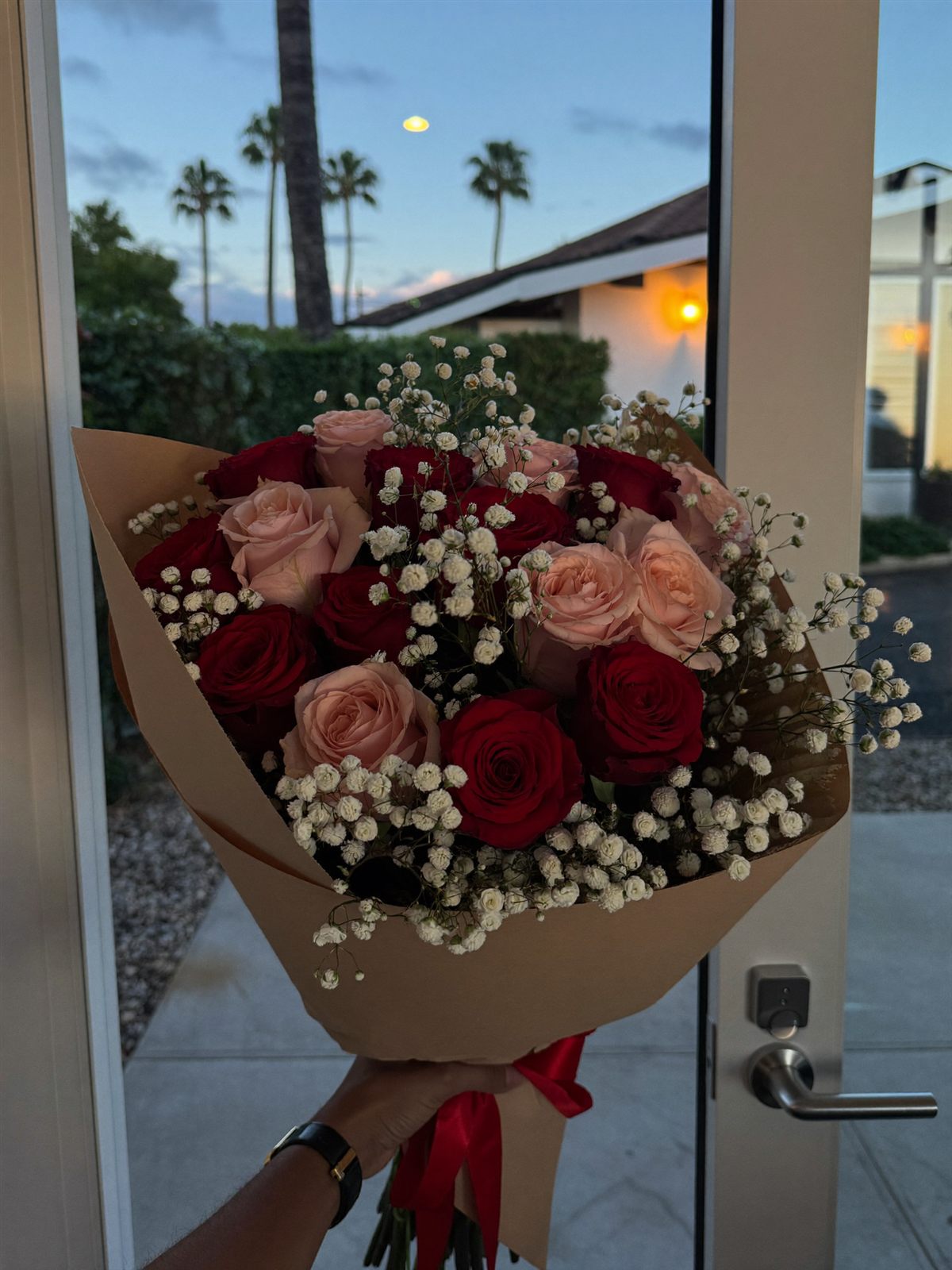 Illustration: red and blush roses with baby's breath, wrapped in kraft paper and held up in a doorway at dusk